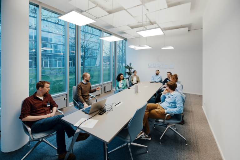 Auf dem Bild sind mehrere Personen in einem Konferenzraum zu sehen, die an einem Workshop teilnehmen. Die Szene vermittelt ein Gefühl von Interaktion und Wissensaustausch in einer professionellen Umgebung.